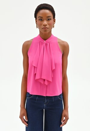 Donna con capelli corti che indossa orecchini ad anello d'oro, una canottiera rosa con dettagli plissettati sul davanti e jeans blu scuro, in piedi contro uno sfondo bianco.