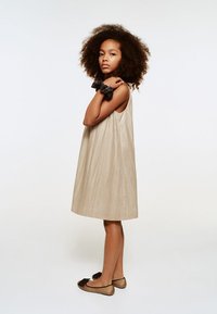 Ljusbeige, ärmlös klänning med en texturerad lyster. Modellen bär ett svart rosettaccessoar och platta skor med bruna detaljer.