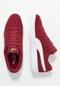 Pár vínově červených semišových tenisek Puma s bílou podrážkou a bočním pruhem, šněrovací design a zlaté logo Puma na jazyku i patě.