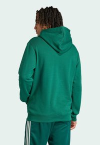 Zelená mikina s kapucňou vyrobená z mäkkej látky, s voľným strihom a rebrovanými manžetami. Viditeľné prešívanie pozdĺž švov a detail kapucne.
