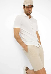 Witte korte mouwen shirt met knopen en zak, gecombineerd met beige shorts en tan sneakers, gedragen met een beige pet.