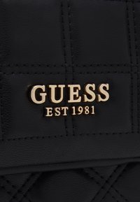 Černý prošívaný kožený povrch s vyraženým zlatým logem "GUESS EST 1981".