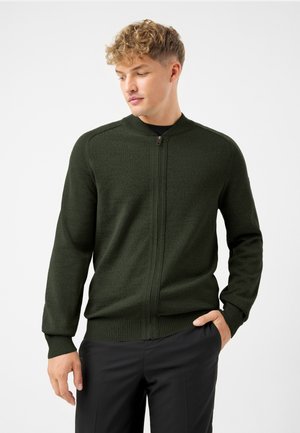 Cardigan zip-up verde scuro realizzato in un tessuto testurizzato, con polsini e orlo a costine e colletto rialzato. Indossato sopra una maglietta nera, esposto su un modello.