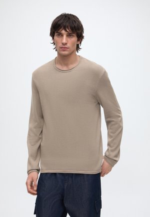Jeune homme aux cheveux foncés portant une chemise beige à manches longues et un pantalon bleu foncé, debout devant un fond clair uni.