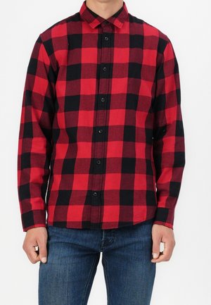 Uomo che indossa una camicia di flanella a quadri rossa e nera abbottonata con maniche lunghe e jeans blu davanti a uno sfondo semplice.