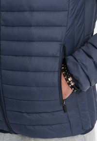 Giacca puffer blu navy con design trapuntato e tasca con zip visibile. Presenta una texture liscia e polsini elasticizzati. Accompagnata da un bracciale di perline.