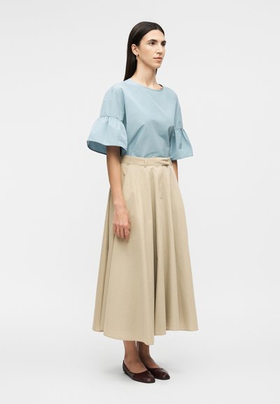 Γυναίκα που στέκεται στραμμένη προς τα δεξιά, φοράει ανοιχτό μπλε μπλούζα με κοντά μανίκια με βολάν, μπεζ μακριά φούστα και σκούρα καφέ flat παπούτσια.