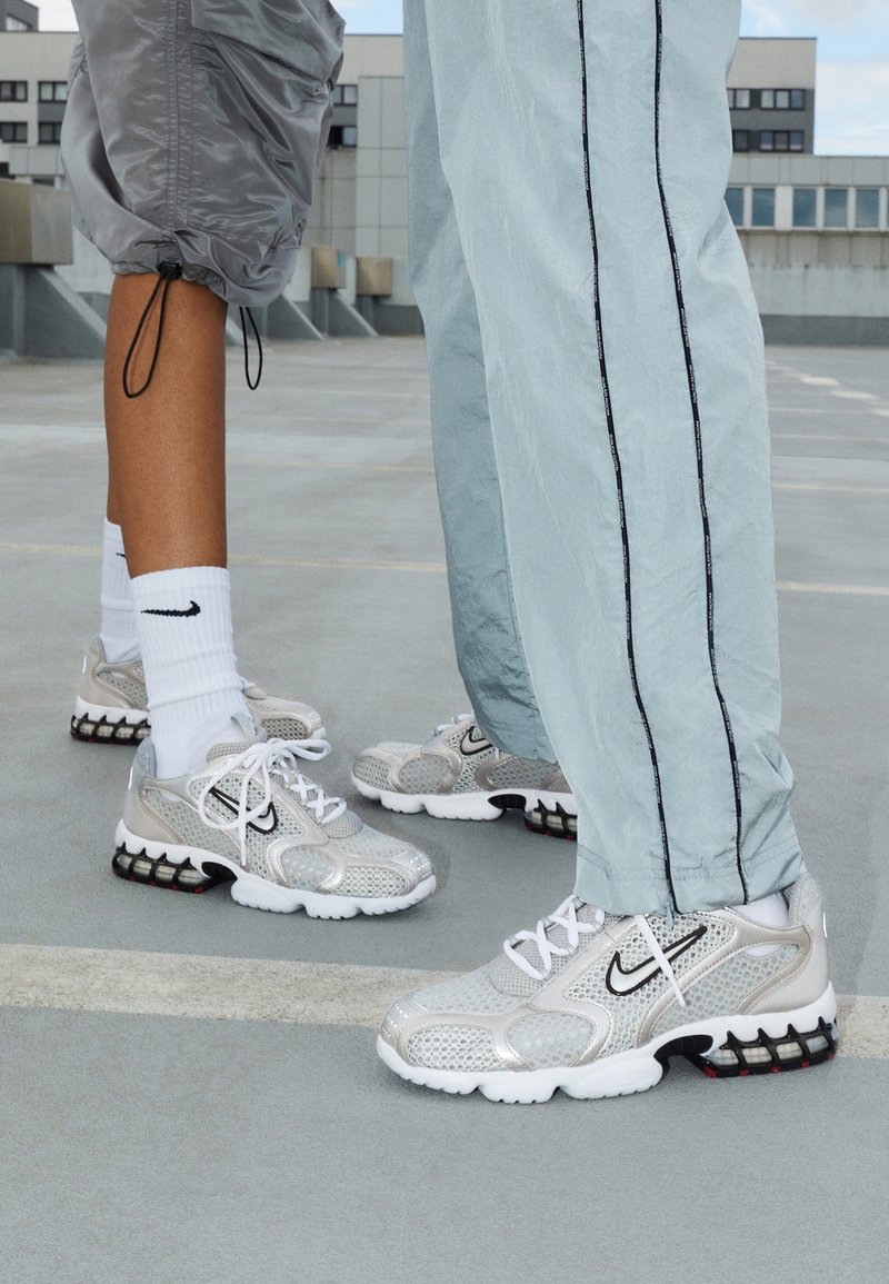 Deux personnes portant des baskets Nike argentées et des chaussettes blanches se tiennent sur une surface en béton avec des bâtiments urbains en arrière-plan.