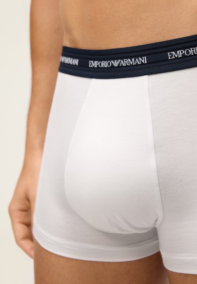 Baltas kokvilnas bokseru apakšveļa ar tumši zilām elastīgām jostām, uz kurām ir "Emporio Armani" logo, gluda tekstūra un pieguloša izstrādājuma dizains.