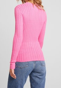 Donna che indossa un maglione rosa a coste, aderente e a maniche lunghe, abbinato a jeans blu, vista da dietro su uno sfondo semplice.