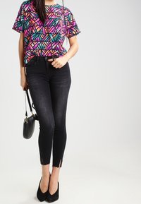 Mujer vistiendo una blusa de manga corta con un colorido patrón geométrico, jeans ajustados negros de cintura alta con aberturas al frente, tacones negros y llevando un bolso negro.