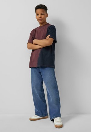 Młoda osoba stoi ze skrzyżowanymi ramionami, ubrana w dwukolorową koszulę bordowo-granatową, niebieskie jeansy z szerokimi nogawkami i białe trampki.