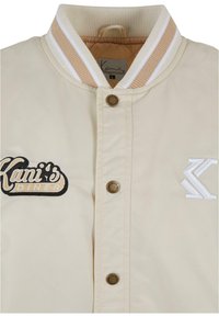 Beige Bomberjacke mit geripptem Kragen in Weiß und Tan, vorderer Knopfschluss und gesticktem "Kani's Diner"-Logo sowie Akzenten.