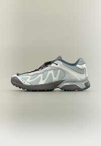 Svetlo sive in bele tekaške superge za tek po poteh z mrežastim zgornjim delom, črnim sistemom hitrega zavezovanja vezalk in robustnim temno sivim podplatom na nevtralni podlagi.