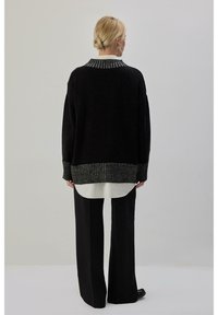 Pull noir surdimensionné avec un col et des poignets côtelés gris texturés, associé à une chemise blanche superposée à l'ourlet, porté avec un pantalon noir.