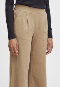 Pantalons beige à jambes larges avec une texture lisse, présentant des plis à l'avant et une taille élastique douce. Un haut sombre à manches longues est visible.