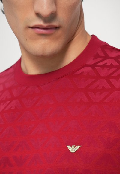 Rdeča majica s kratkimi rokavi, teksturiranim toniranim vzorcem, okroglim izrezom in majhnim zlatim logotipom na levi strani prsnega koša. Gladek zaključek blaga.