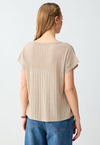 Femme aux longs cheveux blonds portant un pull beige à manches courtes côtelé et un jean en denim bleu, debout dos à un fond uni.