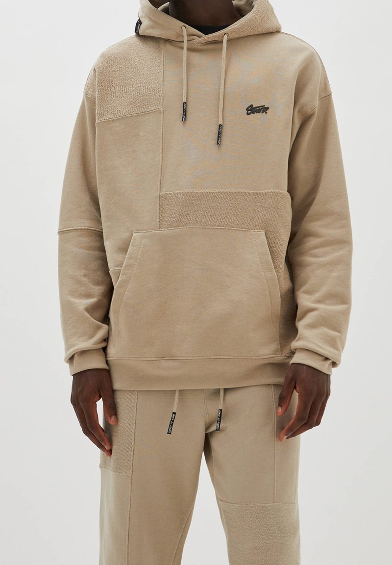 Sweat à capuche beige avec textures variées et patchs, doté d'une poche avant, de cordons de serrage, et d'un petit logo noir sur la poitrine.