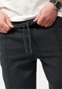 Schwarze Sweatpants mit einem Kordelzugbund, die eine weiche Textur und sichtbare Nahtdetails aufweisen. Eine Nahaufnahme von Händen, die den Bund anpassen.