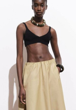 Bralette noir court associé à une jupe taille haute kaki. Le mannequin porte un collier de perles imposant et un bracelet au poignet.