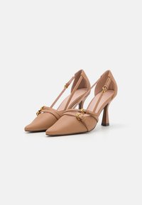 Zapatos de tacón alto de cuero nude con punta afilada, correas en el tobillo y hebillas doradas. Presentan una textura suave y un diseño de tacón esbelto.