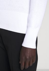 Maglione bianco lavorato a maglia con polsini a coste, texture liscia e vestibilità rilassata, abbinato a pantaloni neri su misura.