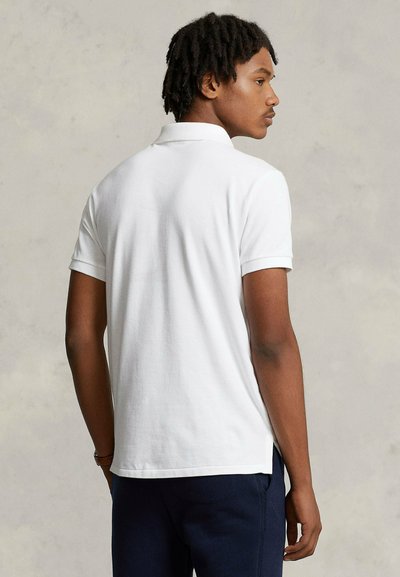 Giovane uomo con dreadlocks corti che indossa una polo bianca semplice a maniche corte e pantaloni scuri, visto di profilo su uno sfondo neutro.