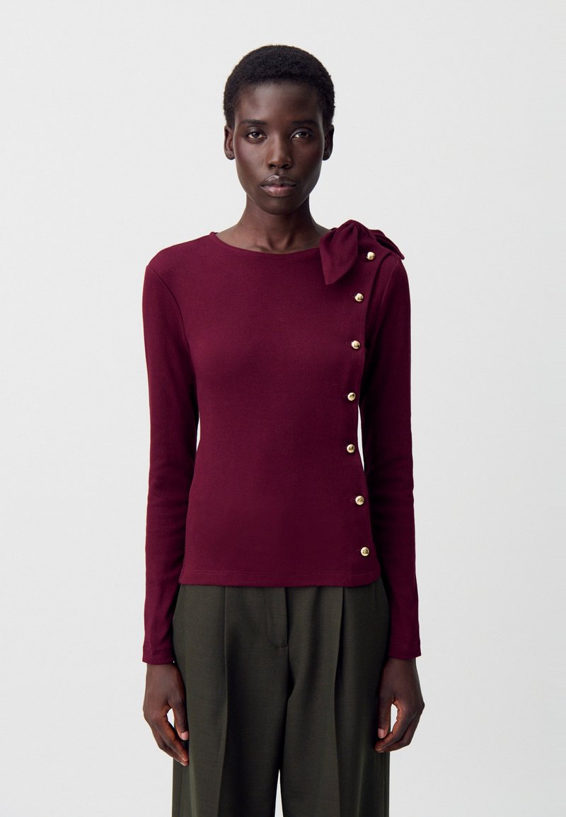 Top lungo bordeaux a maniche lunghe con dettaglio a nodo sulla spalla e bottoni color oro lungo il lato; realizzato in un tessuto morbido e testurizzato.