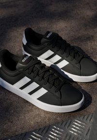 Paire de baskets noires Adidas avec des bandes blanches et des semelles blanches, placées en extérieur sur une surface de sol texturée à la lumière naturelle.
