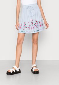 Jasnoniebieska, warstwowa spódnica z haftem kwiatowym w kolorze różowym i zielonym. Posiada elastyczny pas i szczegóły z frędzlami. Połączona z białymi sandałami.