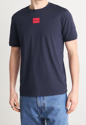 Mann trägt ein dunkelblaues Hugo T-Shirt mit rotem Logo-Patch und hellblauen Jeans, steht mit entspannten Armen an den Seiten.
