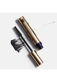 Gullfarget sylindrisk maskarabeholder ved siden av en svart bust-applikator, med svart maskara smurt utover på en hvit overflate. Teksten viser merkenavn og beskrivelse.