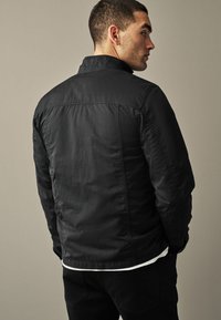 Homme portant une veste noire sur une chemise blanche, de profil, les mains dans les poches devant un fond beige uni.