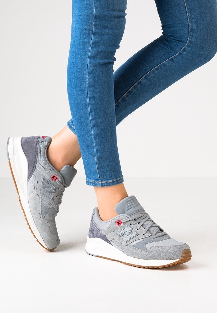 Zapatillas deportivas grises con detalles de ante y malla, que cuentan con una suela de goma, acentos en gris oscuro contrastantes y un logotipo rojo en el costado.