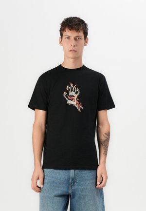 Jonge man in een zwart T-shirt met een afbeelding van een vlammenwerpende skelethand met een mond op de voorkant, gecombineerd met lichtblauwe jeans, staand tegen een witte achtergrond.