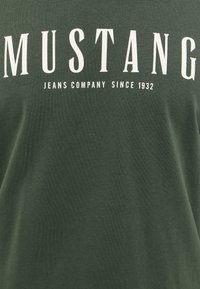 Mörkgrön bomullströja med fet vit text som står "MUSTANG" och "JEANS COMPANY SINCE 1932" i ett avslappnat, modernt teckensnitt.