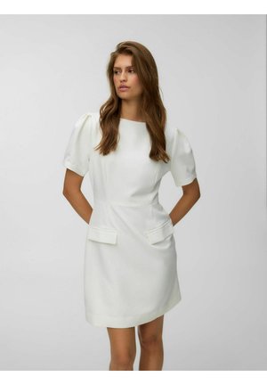 Femme aux longs cheveux bruns portant une robe blanche à manches courtes avec des poches, debout les mains derrière le dos sur un fond uni.