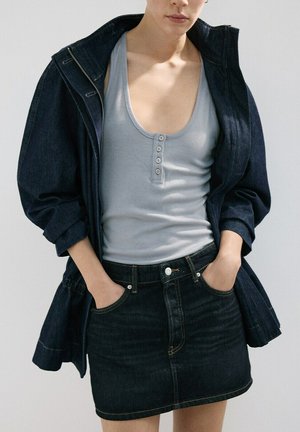 Persona con camiseta de tirantes gris claro de canalé con botones, chaqueta vaquera oscura y falda vaquera oscura, con las manos en los bolsillos.