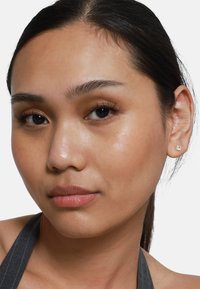Gros plan sur un visage avec une peau lisse ; cheveux bruns tirés en arrière. Petite boucle d'oreille en argent avec un design floral. Maquillage neutre qui met en valeur les traits.