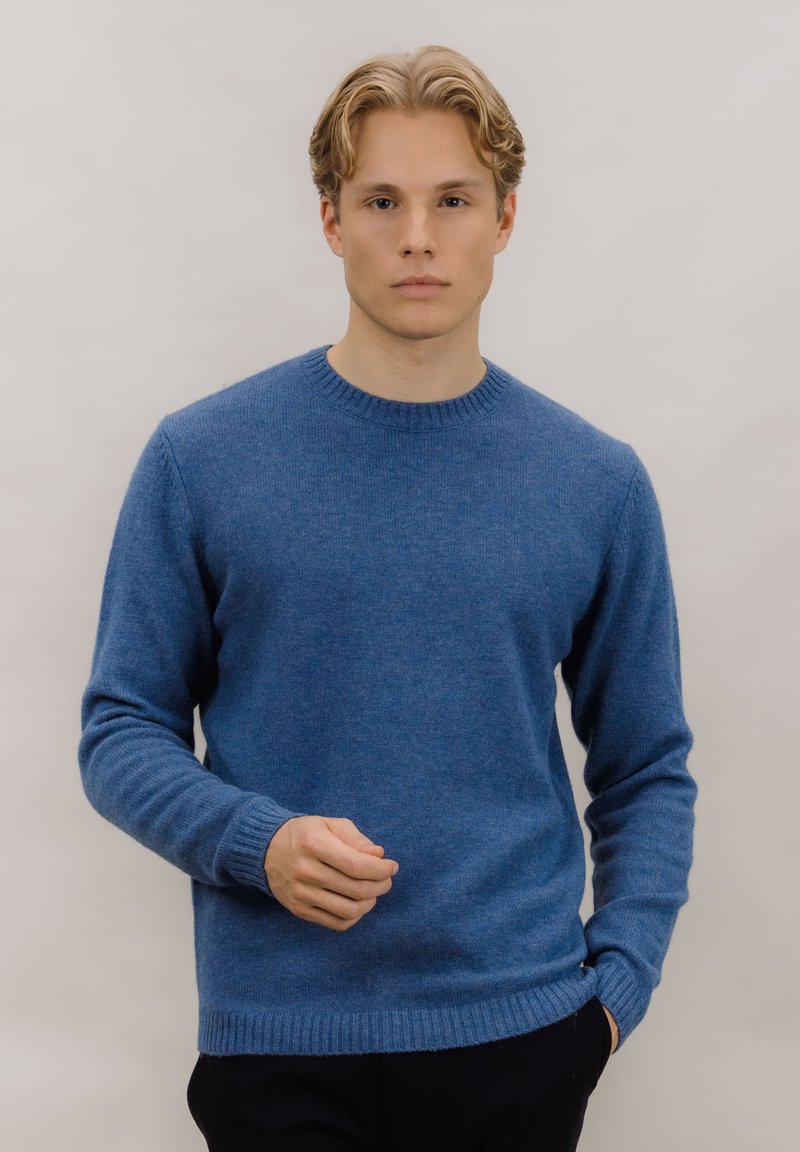 Blå ullgenser med rund hals, ribbestrikkede mansjetter og kant, og en myk tekstur. Modellen står foran en nøytral bakgrunn.