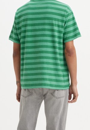 Personne portant une chemise à manches courtes horizontale rayée verte et un jean gris clair, vue de dos sur un fond uni.