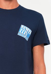 Sötétkék pamut póló, amelyen fehér és világoskék grafikus logó található "tommy JEANS" felirattal a bal mellkas területén. Klasszikus kerek nyakkivágás.