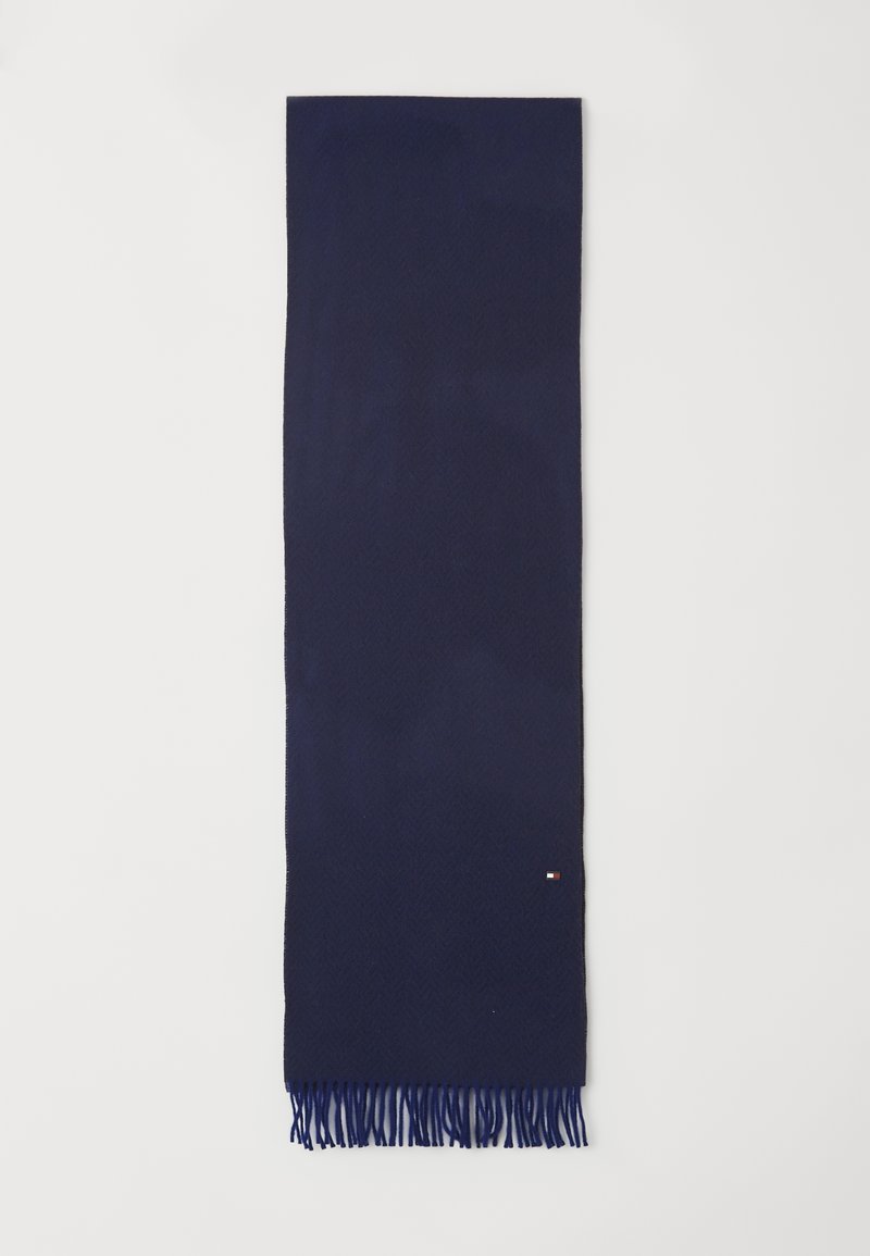 Mørkeblå vævet tørklæde med et fiskebensmønster, afsluttet med frynser i kanterne og et lille logo-tag i den ene ende.