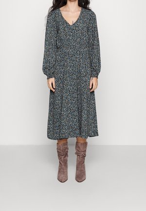 Kvinde iført en langærmet, sort midi-kjole med blomsterprint og lysebrune ruskindsstøvler, stående mod en ensfarvet baggrund.