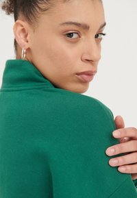 Jonge vrouw met krullend haar in een groene top kijkt over haar schouder, terwijl ze stof aanraakt en zilveren oorbellen in hoepelvorm draagt, tegen een effen achtergrond.