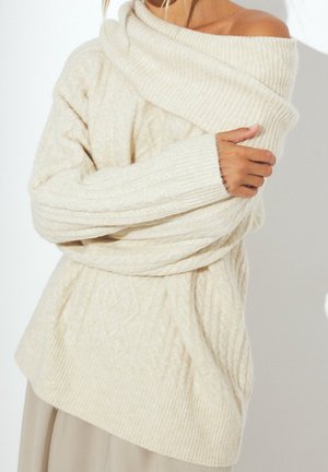 Femme portant un pull en grosse maille torsadée crème à épaules dénudées, les bras croisés, devant un fond clair uni.