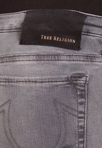 Jeans de mezclilla gris con una etiqueta de cuero negro con "True Religion" en relieve dorado. Incluye costuras detalladas y un diseño en el bolsillo trasero.