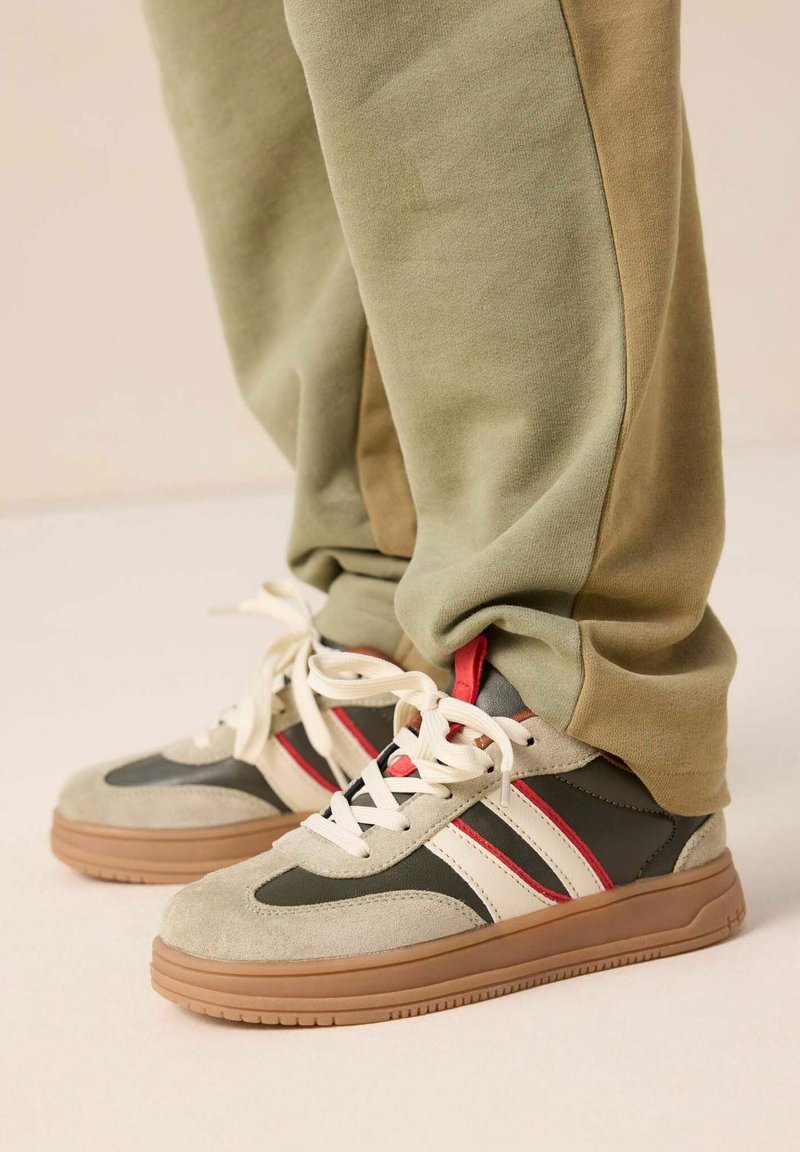 Baskets de sport avec une tige en daim beige agrémentée d'accents vert foncé, de rayures blanches et de détails rouges, associées à un jogging vert clair.