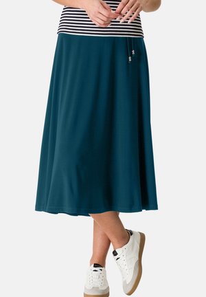Femme portant une jupe midi bleu canard, un haut à rayures et des baskets blanches, debout avec les jambes croisées et les mains jointes devant.